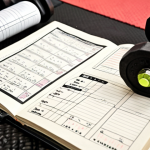 **

"A detailed, close-up shot of a training journal lying open on a tatami mat. The journal page features handwritten notes detailing sets, reps, weights used, and personal sensations during a weightlifting exercise. A pen rests on the page. In the background, a blurred weightlifting rack and gym equipment are visible, suggesting a typical French 'salle de sport'."

**